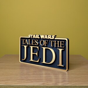 May include: A rectangular sign with the words "STAR WARS TALES OF THE JEDI" in gold lettering on a black background. The sign is made of a dark material and has a gold border. The sign is placed on a wooden surface.