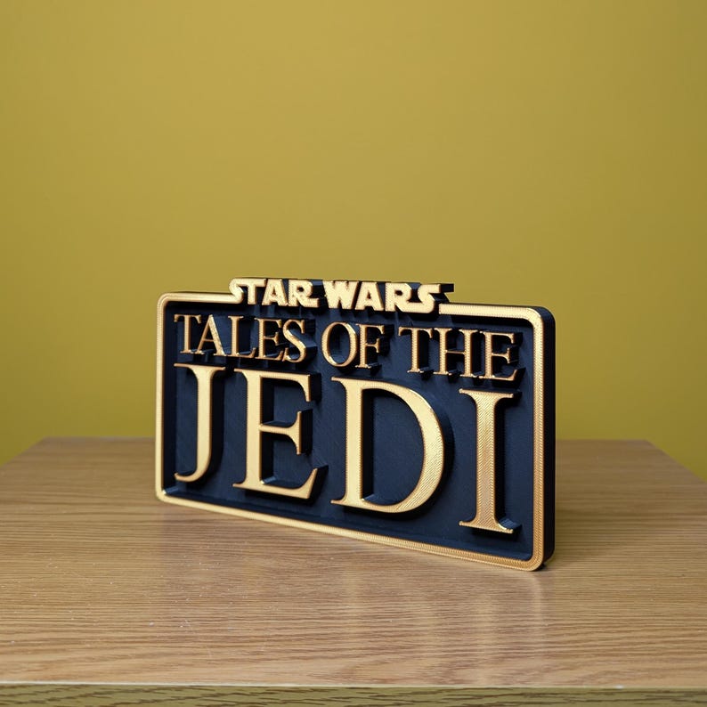 May include: A rectangular sign with the words "STAR WARS TALES OF THE JEDI" in gold lettering on a black background. The sign is made of a dark material and has a gold border. The sign is placed on a wooden surface.