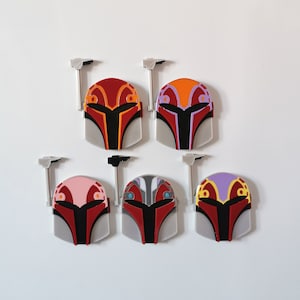 May include: Six Mandalorian helmet charms in different colors. The helmets are red, black, and white with different colored accents. The charms are made of metal and have a hook on the back for attaching to a bag or keychain.