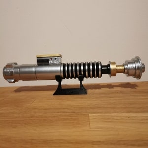 May include: A silver and black lightsaber replica with a gold accent on the handle. The lightsaber is mounted on a black stand.