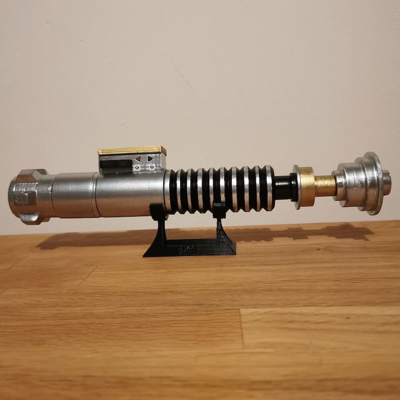 May include: A silver and black lightsaber replica with a gold accent on the handle. The lightsaber is mounted on a black stand.