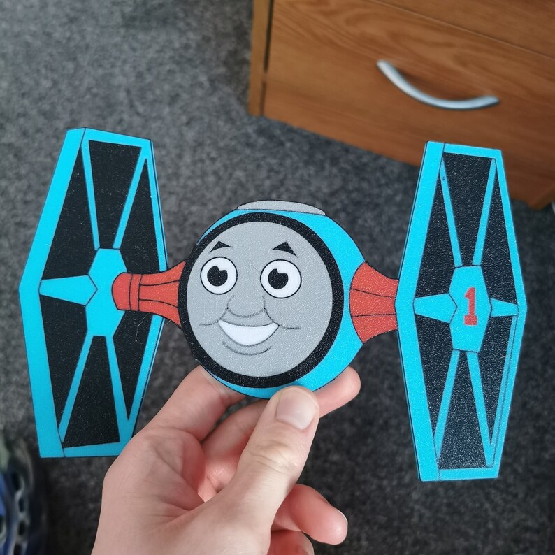 以下が含まれることがあります： トーマス機関車の顔をした青と黒の紙製のTIEファイター。TIEファイターは側面に赤と白のストライプがあり、翼に「1」の数字が書かれている。