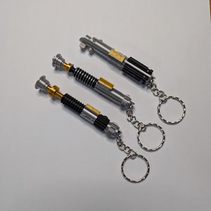 May include: Three silver lightsaber keychains with black, gold, and silver detailing. Each keychain has a silver chain and a silver keyring. The lightsabers are arranged diagonally on a white surface.