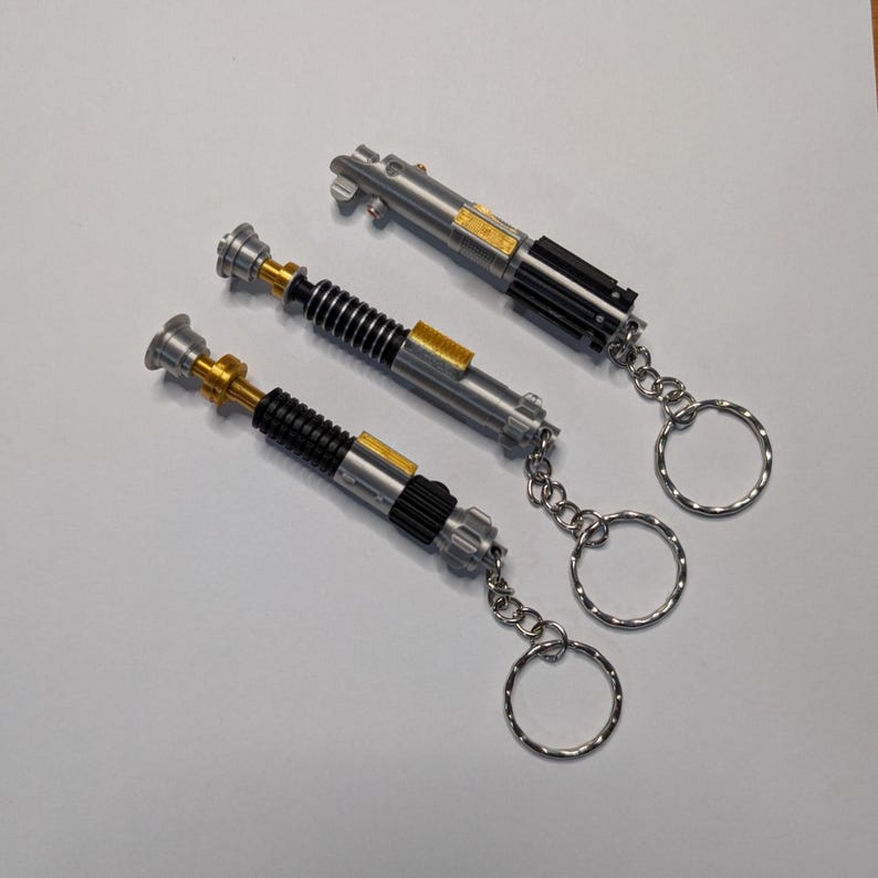 May include: Three silver lightsaber keychains with black, gold, and silver detailing. Each keychain has a silver chain and a silver keyring. The lightsabers are arranged diagonally on a white surface.