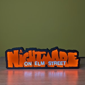 May include: A rectangular, illuminated sign with the text "A Nightmare on Elm Street." The sign features orange lettering with a black outline and a white "ON ELM STREET" text. The sign is lit from within, creating a glowing effect.