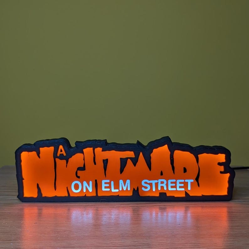 May include: A rectangular, illuminated sign with the text "A Nightmare on Elm Street." The sign features orange lettering with a black outline and a white "ON ELM STREET" text. The sign is lit from within, creating a glowing effect.
