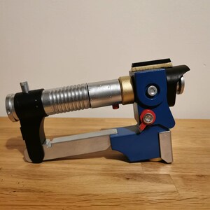 May include: A blue and silver toy blaster with a black barrel and a gold accent. The blaster has a red button and a black handle.