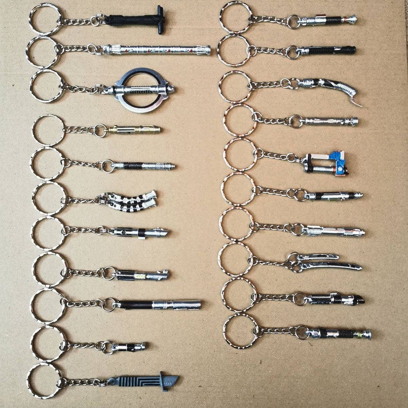 Op de afbeelding: Een verzameling van 18 zilveren sleutelhangers met verschillende Star Wars lightsaber-ontwerpen. Elke sleutelhanger heeft een zilveren ring en een zilveren ketting die is bevestigd aan een miniatuur lightsaber-replica.