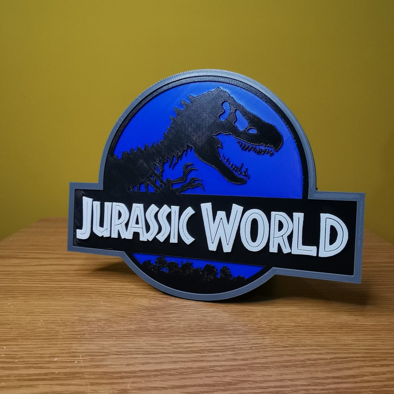 May include: A blue and gray 3D printed sign with the Jurassic World logo. The logo features a silhouette of a dinosaur with the words "Jurassic World" in white text.