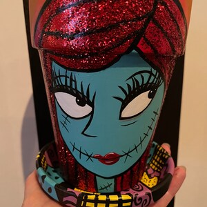 May include: A hand-painted terracotta planter with a red glitter hair and blue face, depicting Sally from the Nightmare Before Christmas. The planter has a matching saucer with a black, yellow, and purple design.