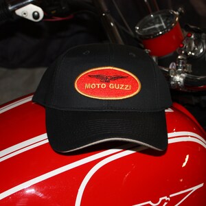 May include: A black baseball cap with a red oval patch that says "MOTO GUZZI" in gold lettering. The patch also features a stylized eagle design.