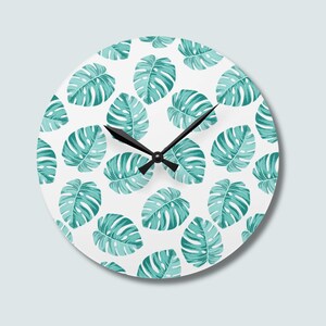 May include: A round wall clock featuring a white background with a repeating pattern of teal monstera leaves. The clock has three black hands positioned at the center, indicating the time, with a clean, circular design and no visible numerals.