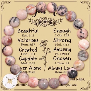 Puede incluir: Una pulsera de cuentas rosas y grises. La pulsera rodea una tarjeta con palabras inspiradoras como "Hermoso" y "Fuerte" y versículos bíblicos. Un pequeño dije de brújula cuelga.