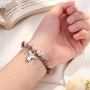 May include: A beaded bracelet with a silver dove charm. The bracelet features round, speckled beads in shades of pink, brown, and cream. The dove charm is detailed with outstretched wings.