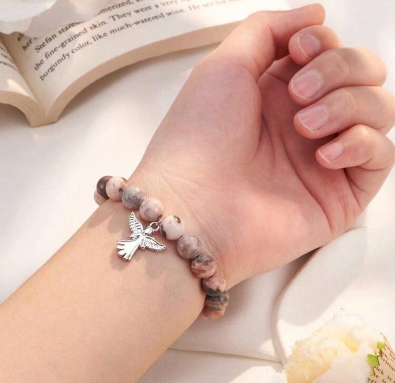 May include: A beaded bracelet with a silver dove charm. The bracelet features round, speckled beads in shades of pink, brown, and cream. The dove charm is detailed with outstretched wings.