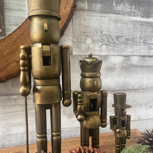 May include: Three gold-colored wooden nutcrackers of varying sizes. The largest nutcracker wears a crown and holds a staff. The other two nutcrackers also wear crowns and hats. The nutcrackers are displayed with pine cones and greenery.