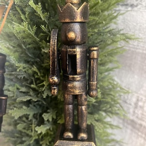 May include: A wooden nutcracker ornament with a crown, shield, and sword. The ornament is painted in shades of brown and gold, with a distressed finish. It is hanging from a gold ribbon, and is a festive holiday decoration.