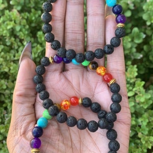 May include: A black lava rock bracelet with seven chakra beads. The beads are red, orange, yellow, green, blue, indigo, and violet. The bracelet is on a person's hand.