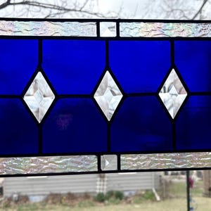 May include: Stained glass window panel with a vibrant blue background and three diamond-shaped clear glass accents. The border features iridescent glass, and the panel is framed in black with hanging hooks.