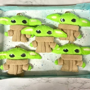 May include: Five felt ornaments shaped like the character Baby Yoda from the Star Wars series. The ornaments are green with large eyes and are wearing brown felt coats.