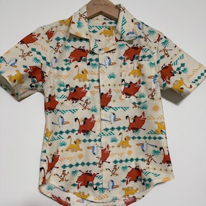 May include: Off-white short-sleeved button-up shirt featuring a repeating pattern of cartoon characters, including lions, warthogs, and birds. The shirt has a collared neckline and two small chest pockets. The shirt is made of a lightweight fabric.