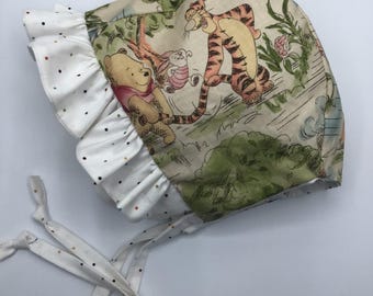 May include: A white bonnet with a Winnie the Pooh and Tigger print. The bonnet has a white ruffle with black polka dots and white ties.