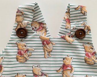 May include: Close-up of a fabric with a Winnie the Pooh pattern. The fabric features a repeating pattern of Winnie the Pooh characters in various poses, set against a white background with green stripes. Two brown wooden buttons are visible.