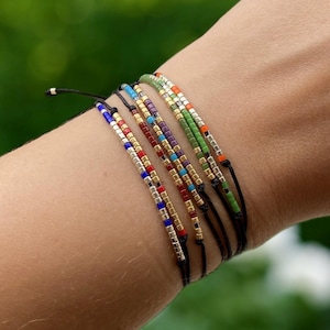 May include: A collection of colorful beaded bracelets on a wrist. Each bracelet features small, square beads in various colors, including red, blue, gold, and green, strung on a black cord. The background is a soft, blurred green.