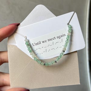 Könnte beinhalten: Ein Perlenarmband mit kleinen, durchscheinenden hellgrünen und klaren Perlen sowie silberfarbenen Zwischenelementen. Das Armband ist auf einer weißen Karte in einem beigen Umschlag befestigt, bedruckt mit "Until we meet again" und Morsezeichen.