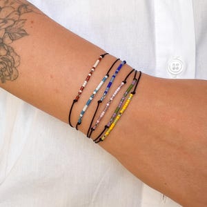 Op de afbeelding: Een verzameling van zes armbanden met kralen in verschillende kleuren, waaronder rood, blauw en geel, geregen op zwarte koorden. De armbanden worden op een arm gedragen, tegen een witte shirt achtergrond. De kralen zijn klein en kleurrijk.