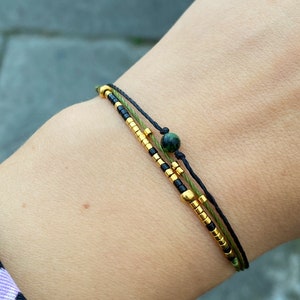 以下が含まれることがあります： 黒、緑、金と黒のビーズが交互に並んだ3本の糸でできたブレスレット。黒い糸に緑色の石のビーズが付いている。