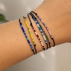 May include: Five thin black cord bracelets, each featuring a unique pattern of small, colourful glass seed beads. The beads are arranged in various sequences of blue, yellow, red, white, green, and purple, creating distinct linear designs.