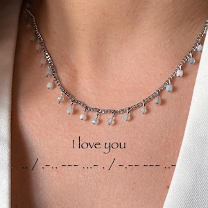 Könnte beinhalten: Eine silberne Halskette mit kleinen, herabhängenden, hellfarbigen Perlen. Die Halskette wird von einer Person getragen, die einen weißen Blazer trägt. Der Text "I love you" und Morsecode sind auf der Brust zu sehen.