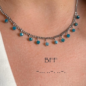 May include: A silver necklace featuring small turquoise beads suspended from the chain. The necklace is worn by a person in a white blazer. The letters "BFF" are printed below the necklace.