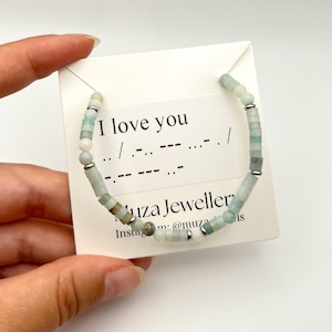 May include: A beaded bracelet featuring light blue, pale green, and white cylindrical beads interspersed with small silver-toned metal spacers. The bracelet is displayed on a white card printed with the text I love you and Morse code.