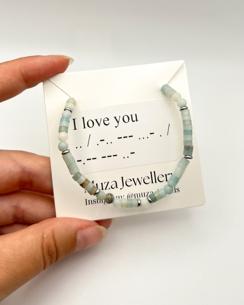 May include: A beaded bracelet featuring light blue, pale green, and white cylindrical beads interspersed with small silver-toned metal spacers. The bracelet is displayed on a white card printed with the text I love you and Morse code.