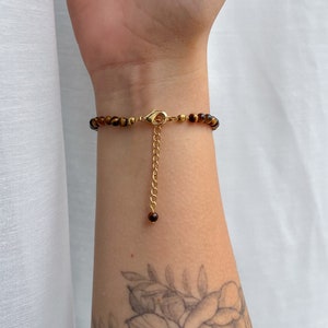 May include: A bracelet featuring small, round, brown and gold tiger eye beads. The piece includes a gold-toned metal lobster claw clasp and a dangling gold-toned chain extender that ends with a single, small, round tiger eye bead accent.