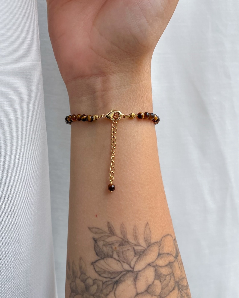May include: A bracelet featuring small, round, brown and gold tiger eye beads. The piece includes a gold-toned metal lobster claw clasp and a dangling gold-toned chain extender that ends with a single, small, round tiger eye bead accent.