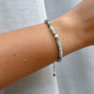 May include: A delicate bracelet featuring a sequence of small, smooth beads in shades of pale green, white, and grey, interspersed with tiny silver-toned metal spacers. The bracelet includes a thin silver-toned chain extender with a single bead.