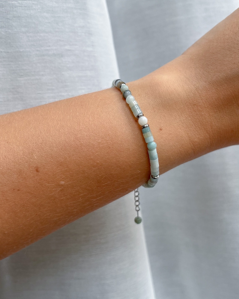 May include: A delicate bracelet featuring a sequence of small, smooth beads in shades of pale green, white, and grey, interspersed with tiny silver-toned metal spacers. The bracelet includes a thin silver-toned chain extender with a single bead.