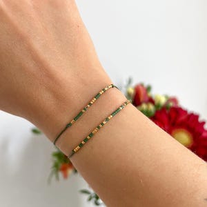 Può includere: Due braccialetti delicati realizzati con un sottile cordino verde scuro, ciascuno caratterizzato da una sequenza di piccole perline color oro metallizzato e verde scuro disposte in un motivo lineare. I braccialetti sono indossati insieme.