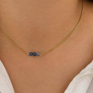 May include: A gold-toned chain necklace featuring three small, round beads in varying shades of blue. The beads are positioned off-center along the delicate metal chain, which has a classic curb link design and a polished, reflective finish.