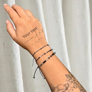 Könnte beinhalten: Drei verstellbare Kordelarmbänder mit kleinen metallischen und schwarzen Perlen, die in Morsecode-Mustern angeordnet sind. Die Armbänder werden am Handgelenk getragen, wobei der Text "Your text" und Morsezeichen auf der Haut über dem Schmuck sichtbar sind.