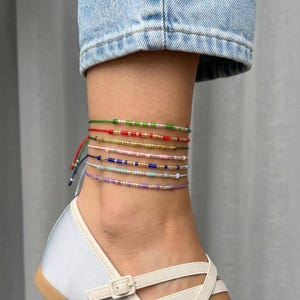 May include: A selection of colourful beaded ankle bracelets. The bracelets, in shades of green, red, pink, blue, and purple, are adorned with small beads and tied with string. They are worn on an ankle, paired with a light-coloured shoe.