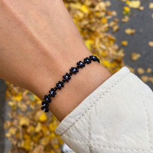 Op de afbeelding: Een zwarte kralen armband met kleine bloemvormige kralen. De armband zit om de pols van een persoon.