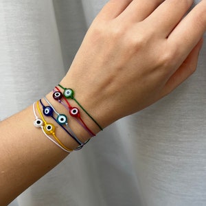 Puede incluir: Un conjunto de cinco pulseras de cordón fino, cada una con una cuenta de vidrio en forma de ojo turco. Las pulseras son de color blanco, amarillo, azul, morado y verde, con cordones a juego. Cada cuenta muestra una pupila negra central.