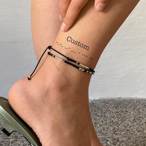 Peut inclure: Bracelet de cheville avec cordon noir et perles argentées. Le bracelet est porté sur la cheville, complété par une chaussure en satin vert. Le mot "Custom" est imprimé sur la cheville.