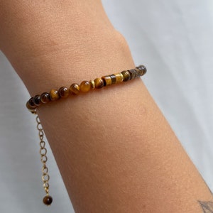 May include: A bracelet featuring small, round, and cylindrical tiger's eye beads in shades of brown and gold, accented with thin gold-toned metal spacers. The piece includes a gold-toned metal chain extender with a single bead at the end.