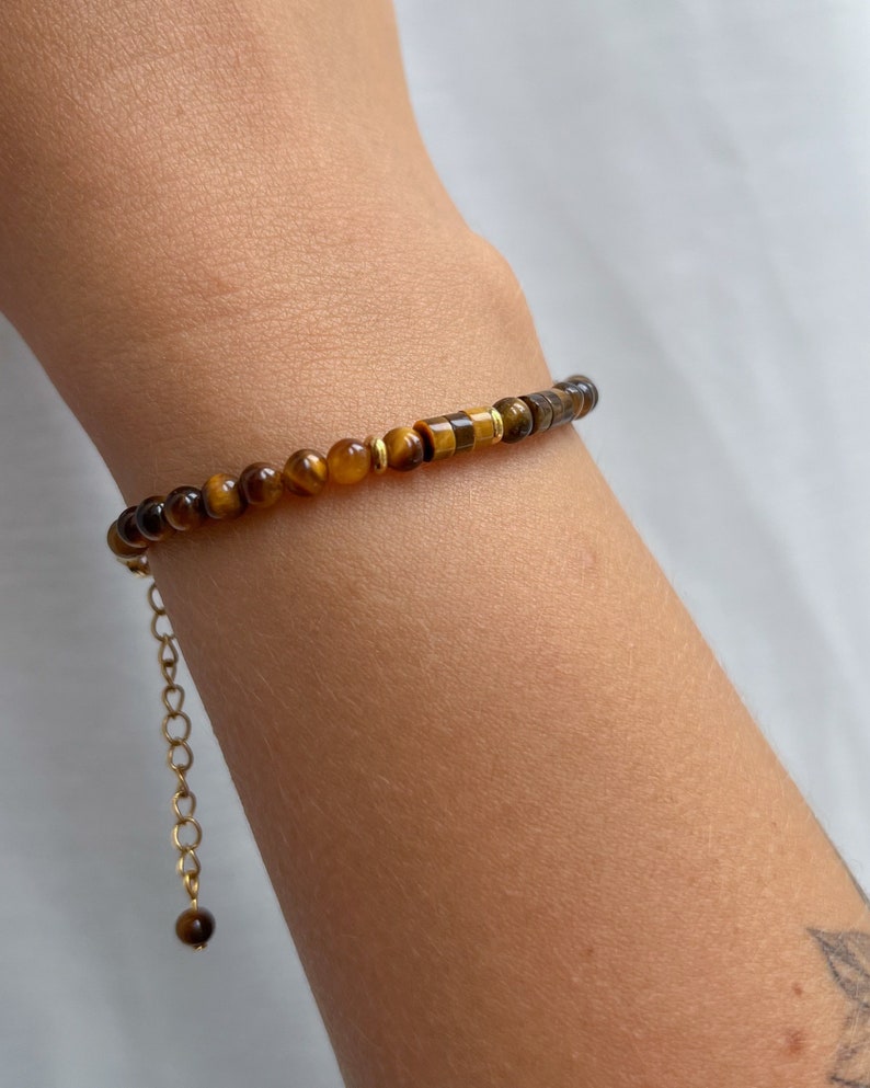May include: A bracelet featuring small, round, and cylindrical tiger's eye beads in shades of brown and gold, accented with thin gold-toned metal spacers. The piece includes a gold-toned metal chain extender with a single bead at the end.