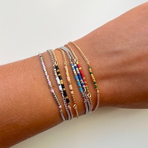 May include: A set of six delicate bracelets featuring thin metallic chains and small, colourful seed beads. The beads are arranged in distinct patterns of pink, white, black, beige, blue, red, and green, with gold and silver-toned chain links.